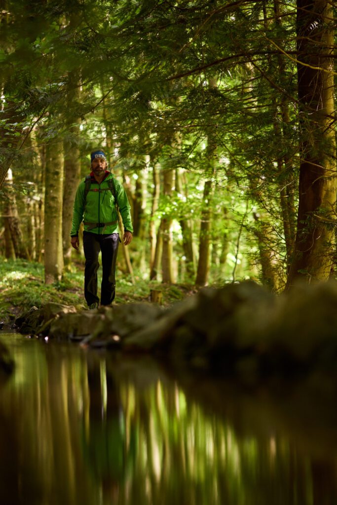 Wanderer mit Grüner Jacke läuft durch Wald mit Fluss
