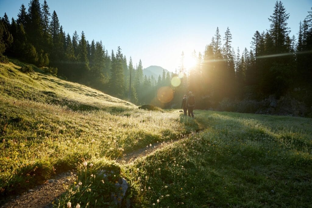 Tourismusbild Österreich Wanderpaar läuft Pfad entlang auf Grüner Wiese mit Wald bei Sonnenaufgang